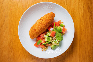 A golden-brown croquette sits next to a colorful salad of tomatoes, greens, and nuts
