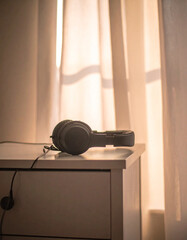 A pair of headphones resting on a small drawer with soft glowing light coming through curtains.