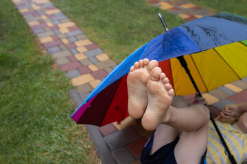 The child's bare feet are raised up and peeking out from under colorful umbrella. Joy, positive,...