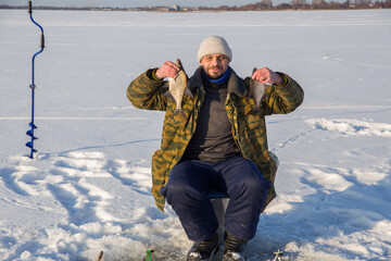 A male fisherman sits on the ice of a lake and holds a fish he caught in his hands. He looks at the camera.