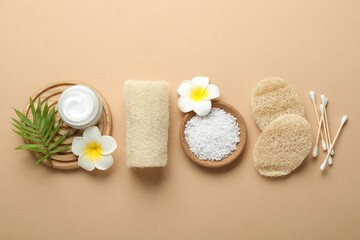 Natural loofah sponges, cosmetic product, sea salt, swabs, plumeria flowers and green twig on beige background, flat lay