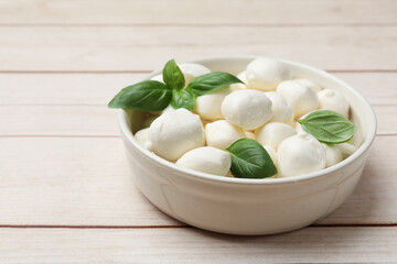 Tasty mozzarella cheese balls in bowl and basil on light wooden table, closeup. Space for text