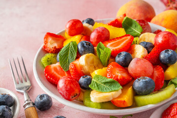 Close up of summer colorful fruit salad in a bowl on pink background. Healthy vegan food with vitamins. Mixed strawberries, grapes, banana, kiwi, blueberries, peach, mint for diet lunch