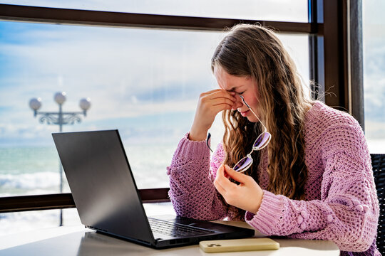 Young woman touching nose bridge and squinting while using laptop in cafe. Eye strain, poor vision, headache from screen exposure, digital fatigue, computer vision syndrome, screen stress indoors