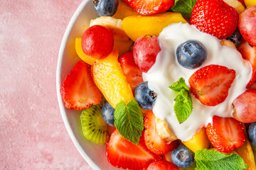 Close up of summer colorful fruit salad with creamy yogurt in a bowl on pink background. Top view. Healthy vegan food with vitamin. Mixed strawberries, grapes, banana, kiwi, blueberries, peach, mint