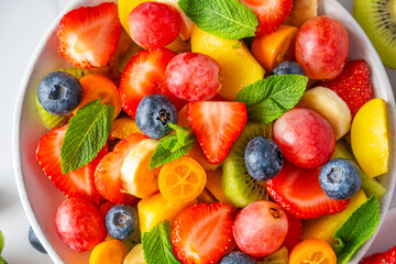 Close up of summer colorful fruit salad in a bowl on white marble background. Top view. Healthy vegan food with vitamin. Mixed strawberries, grapes, banana, kiwi, blueberries, peach, citrus