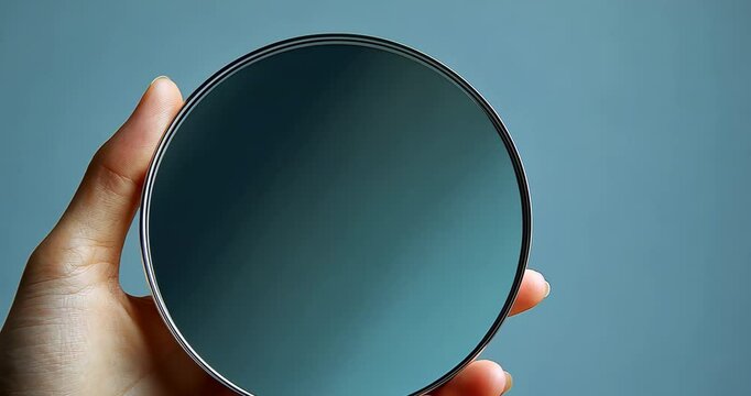 A hand holds a round mirror against a light blue background. The mirror reflects light and shadow, creating a gradient effect.