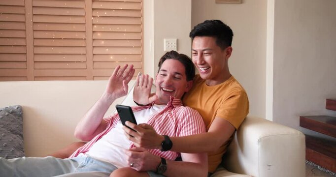 Happy diverse gay couple relaxing on couch, smiling and watching video on smartphone, at home