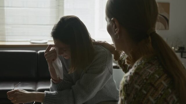 Two women in therapy circle, one visibly emotional and wiping tears with tissue, the other gently placing hand on her shoulder, showing empathy and emotional support in softly lit room
