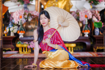 Elegant woman in traditional Thai Chakkraphat costume holding a paper umbrella, seated in a temple setting.