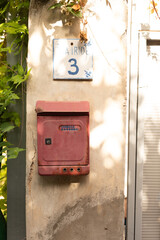 A vintage red mailbox on an aged wall, next to a house number plaque.  Perfect for rustic, vintage, or travel themes.