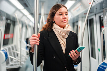 A young woman is engaged in using her smartphone while traveling on the subway, holding the pole with one hand and browsing various applications with her other hand, fully immersed in her mobile