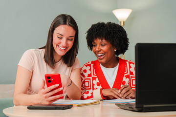 Two cheerful women enjoying a lively interaction as they use a smartphone, sharing laughter and...