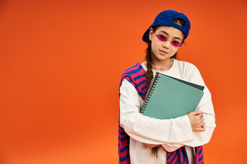 Stylish teenage girl poses in trendy outfit against vibrant orange backdrop in studio