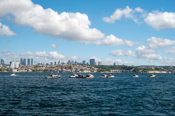 Fototapeta premium Fishing boats on the Bosphorus with large ships passing under the Bosphorus Bridge in Istanbul, Turkey on a bright day with scenic clouds, hills, and a Turkish flag in the background.