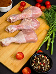 Top view arrangement of raw chicken legs, cherry tomatoes, dill and seasonings: paprika, pepper and salt on wooden board neatly arranged on dark background, demonstrating fresh culinary delights