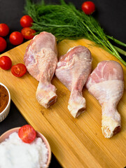 Top view arrangement of raw chicken legs, cherry tomatoes, dill and seasonings: paprika, pepper and salt on wooden board neatly arranged on dark background, demonstrating fresh culinary delights