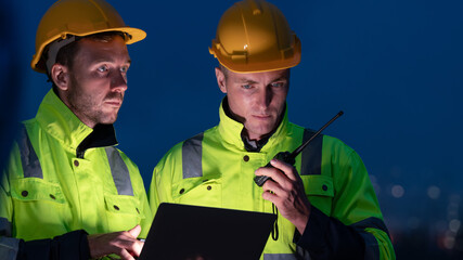 caucasian white engineers in safety uniforms and helmets using laptop and radio for site communication. Concept of teamwork, inspection, and operations at an industrial facility.Construction of Oil, 