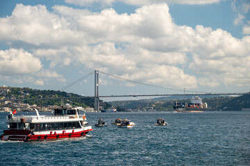 Fishing boats on the Bosphorus with large ships passing under the Bosphorus Bridge in Istanbul, Turkey on a bright day with scenic clouds, hills, and a Turkish flag in the background.