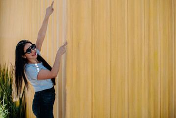 Fototapeta premium Trendy woman wearing sunglasses, smiling and pointing upward, yellow background enhances positivity.
