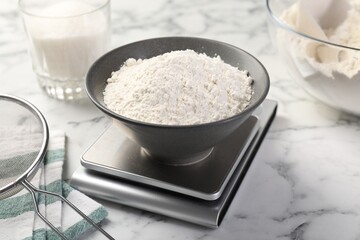 Digital kitchen scale with bowl of flour and other products on white marble table, closeup