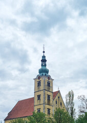 The tall, ornate steeple of St. Anastasia Church with a red-tiled roof stands prominently against a cloudy sky in Samobor