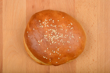 A single, golden brown burger bun sprinkled with sesame seeds sits on a light wooden surface