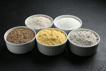 Different types of flour in bowls on black textured table, closeup