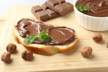 Tasty toast with chocolate spread, hazelnuts and mint on wooden board, closeup