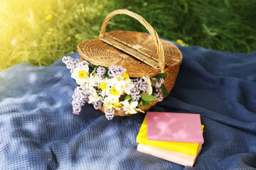 Stacked books, blanket and wicker basket with flowers on green grass outdoors