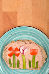 A slice of bread spread with pate is decorated with vegetables to resemble flowers on a blue plate