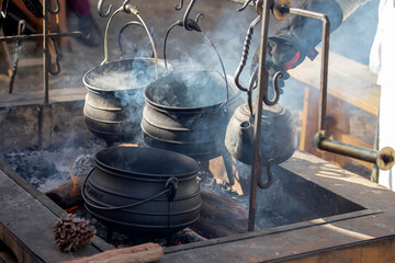 Traditional cast iron pots and kettle hanging over an open fire pit with rising smoke at outdoor historical camp. 