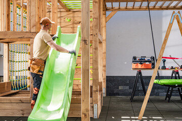 Construction Worker Installs Green Slide in Wooden Playset During Bright Daylight at a Residential Backyard