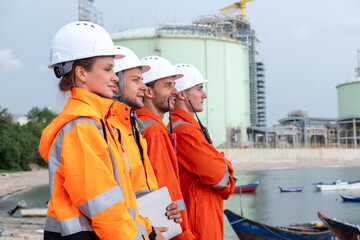 Workers in safety gear observe operations at an industrial site near the water during daytime