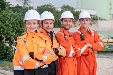 Team of workers in bright orange uniforms and helmets pose confidently near the water at a construction site in the evening