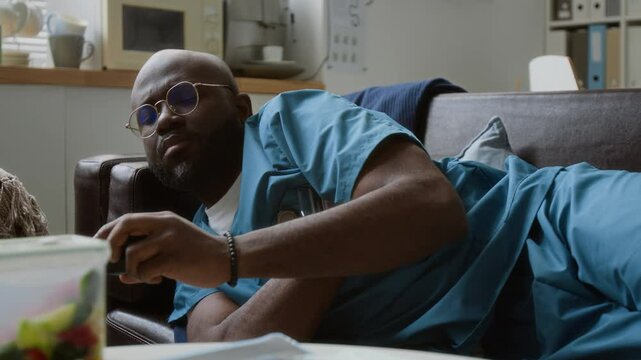 Sleepy African American male clinician in medical scrubs napping on couch in break room, taking mobile gadget from table, getting up and hurrying to sick patient