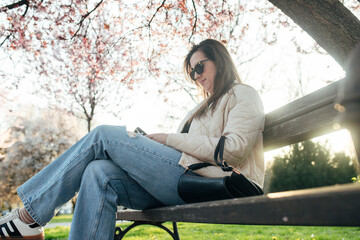 Woman enjoying a sunny day in the park while using her phone on a bench surrounded by cherry...