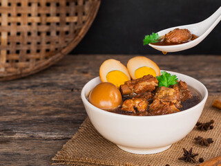 Use spoon to pick up stewed pork ribs and boiled egg in sweet brown sauce in bowl on wooden background. Thai Food
