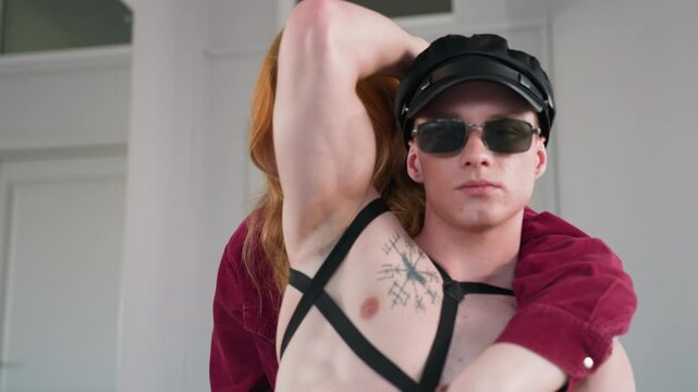 front view of man wearing harness cap and sunglasses seated in dance studio beside mirror as partner embraces from behind cuffed hands between them creating intimate playful mood