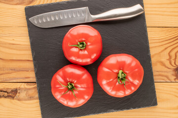 Three pink tomatoes with slate tray and knife on wooden table, close-up, top view.
