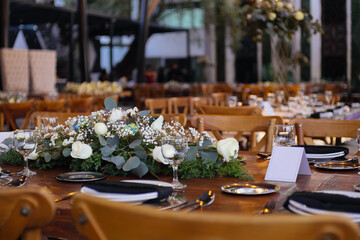 A long wooden table is set for a formal event with white dishes, black napkins, glassware, and a centerpiece of white roses and greenery. The sophisticated arrangement is perfect for weddings