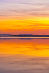 Vivid sunset over a calm lake with golden and purple skies mirrored in the water. A tranquil, dreamlike scene full of color and serenity.