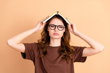 Young woman with glasses holding a book overhead looking displeased against a beige background