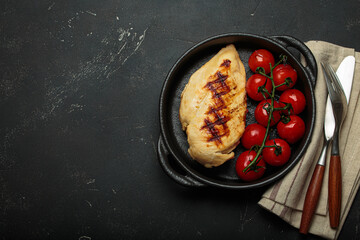 Grilled chicken breast in a cast iron pan on rustic black background, surrounded by roasted cherry tomatoes and cutlery, healthy meal concept, copy space