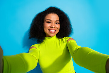 Young woman in neon green top smiling and taking a selfie against a vibrant blue background...