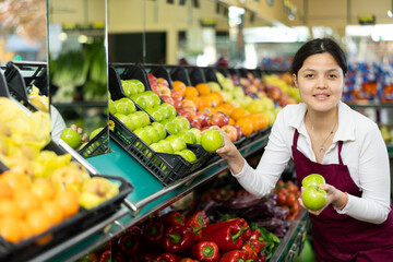 Young woman in apron working in salesroom of greengrocer, setting out fruits on shelves