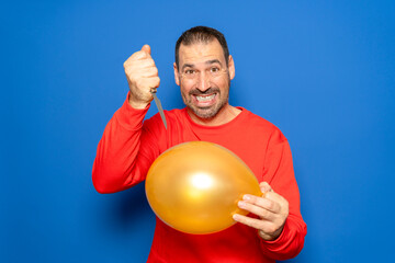 Hispanic man with a beard in his 40s wearing a red sweater about to burst a balloon with a knife with the intention of surprising someone, isolated on a blue studio background.
