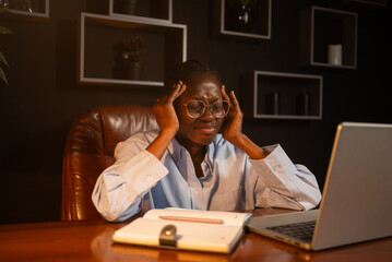Young african businesswoman struggling with a severe headache while working late at night in the office, massaging her temples in an attempt to ease the discomfort and tension