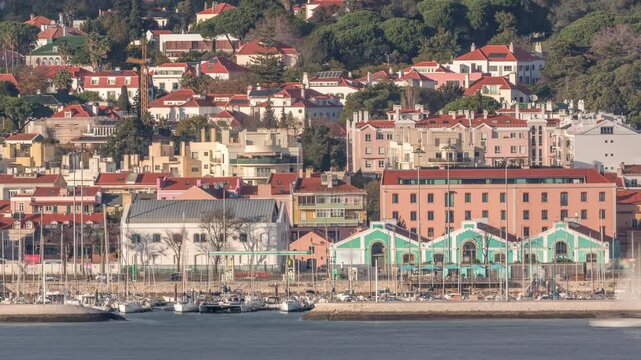 Aerial timelapse of Marina Doca do Bom Sucesso in Lisbon, Portugal, once a naval harbor and now a leisure port. Colorful houses with red roofs line the waterfront against the Tagus River backdrop.