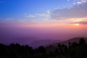 Mountain view at sunrise or sunset, with a purple and blue gradient sky in the background and shadows of trees in the foreground from the low light in the morning or evening.